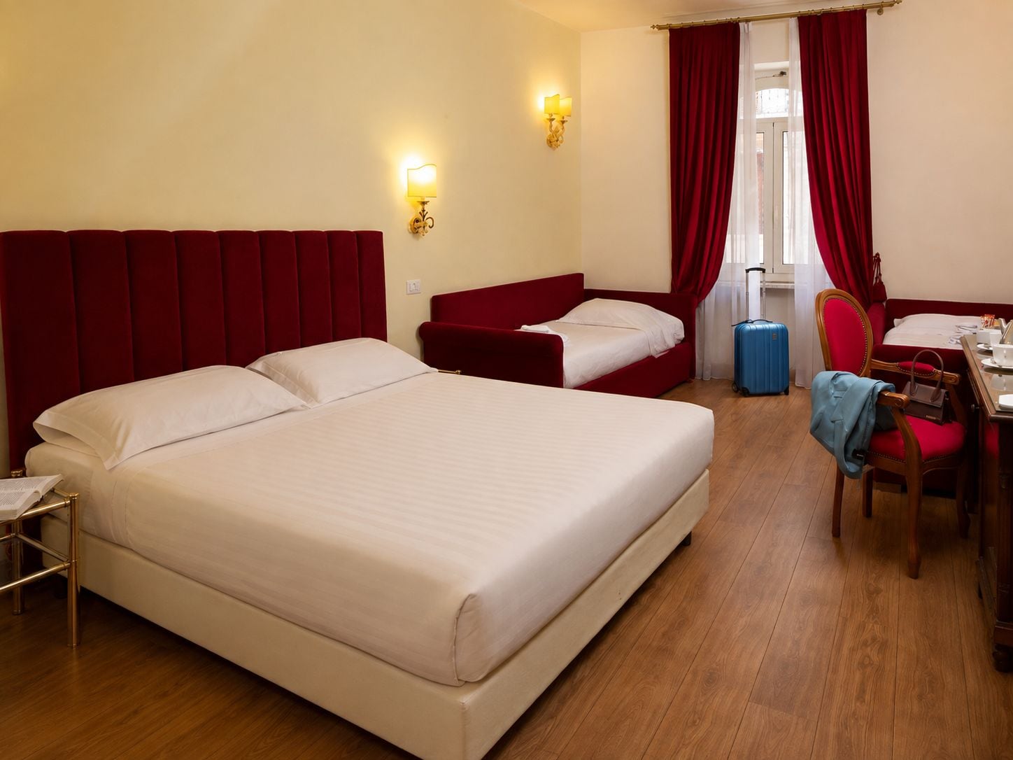 A hotel room with a large bed, a smaller bed, a desk with a chair, a suitcase, and a window with red curtains.