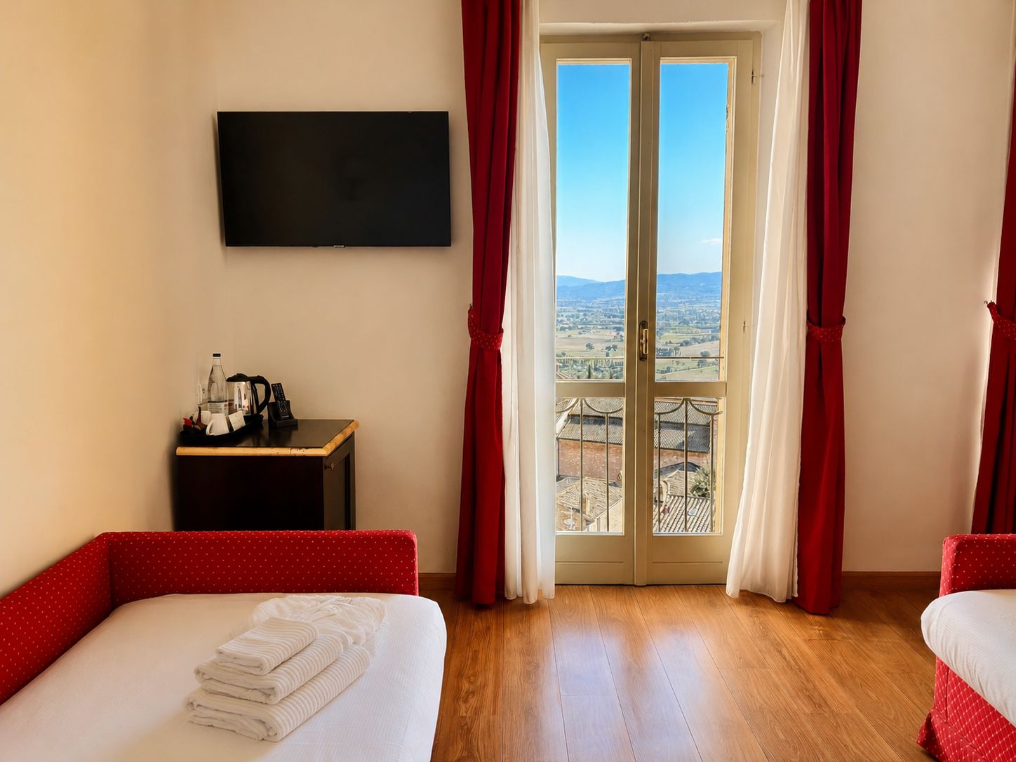 Habitación de hotel con cama, televisión, una mesita con teléfono y botella, y un balcón con cortinas rojas.