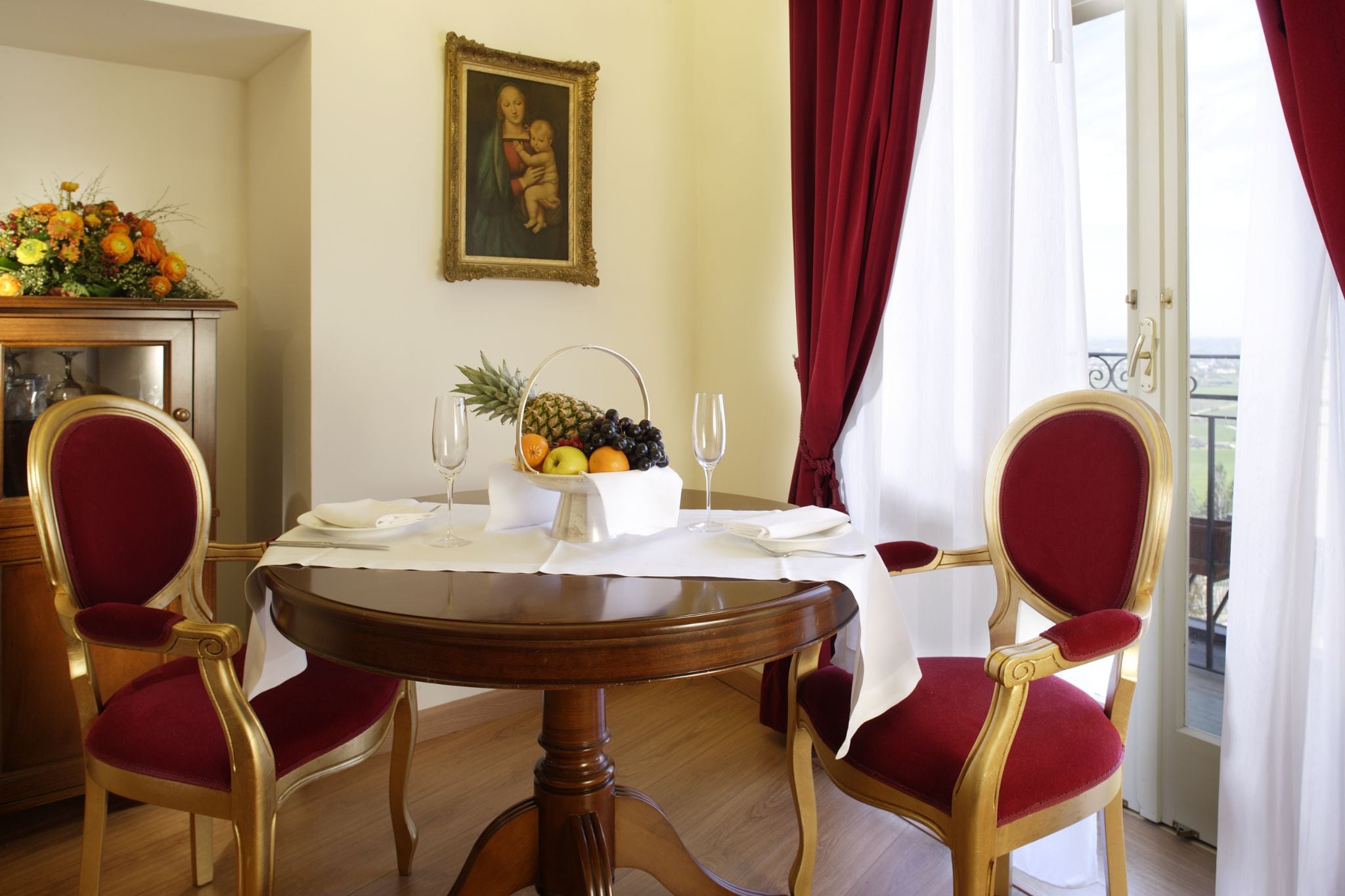 Een eetkamer met een ronde tafel gedekt voor twee personen, twee stoelen, een fruitmand en een schilderij aan de muur.