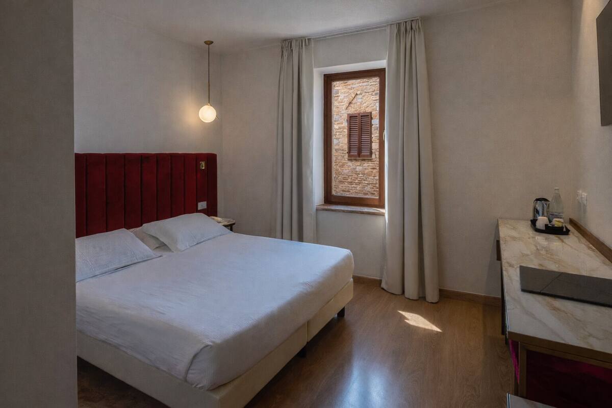 A hotel room with a bed, a nightstand, a window with curtains, and a small table with a lamp.