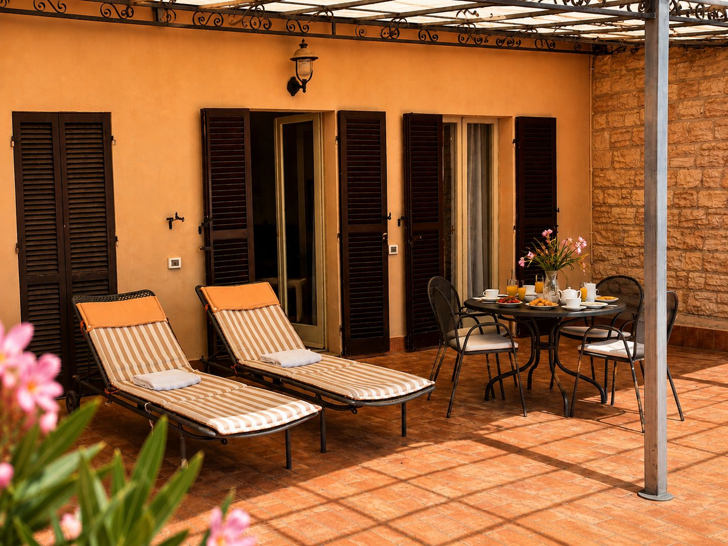 A sunlit patio with two striped lounge chairs, a table with chairs, and a stone wall.