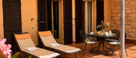 A sunlit patio with two striped lounge chairs, a table with chairs, and a stone wall.