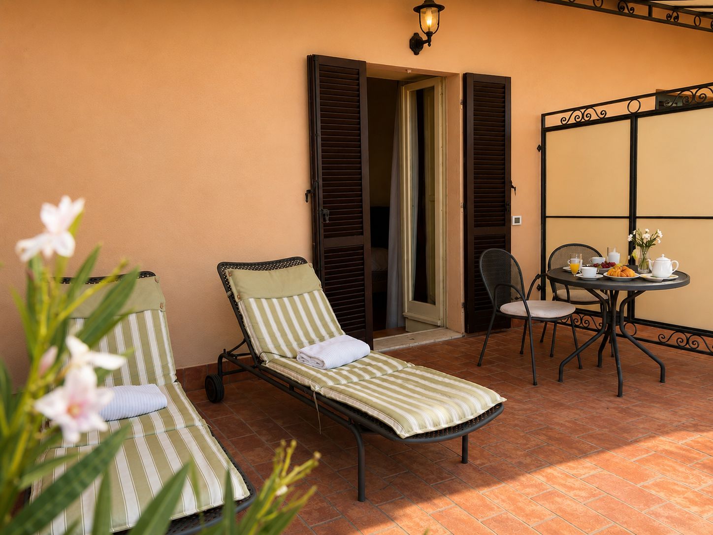 A sunlit outdoor seating area with two striped lounge chairs, a table with breakfast items, and a door leading to an interior space.