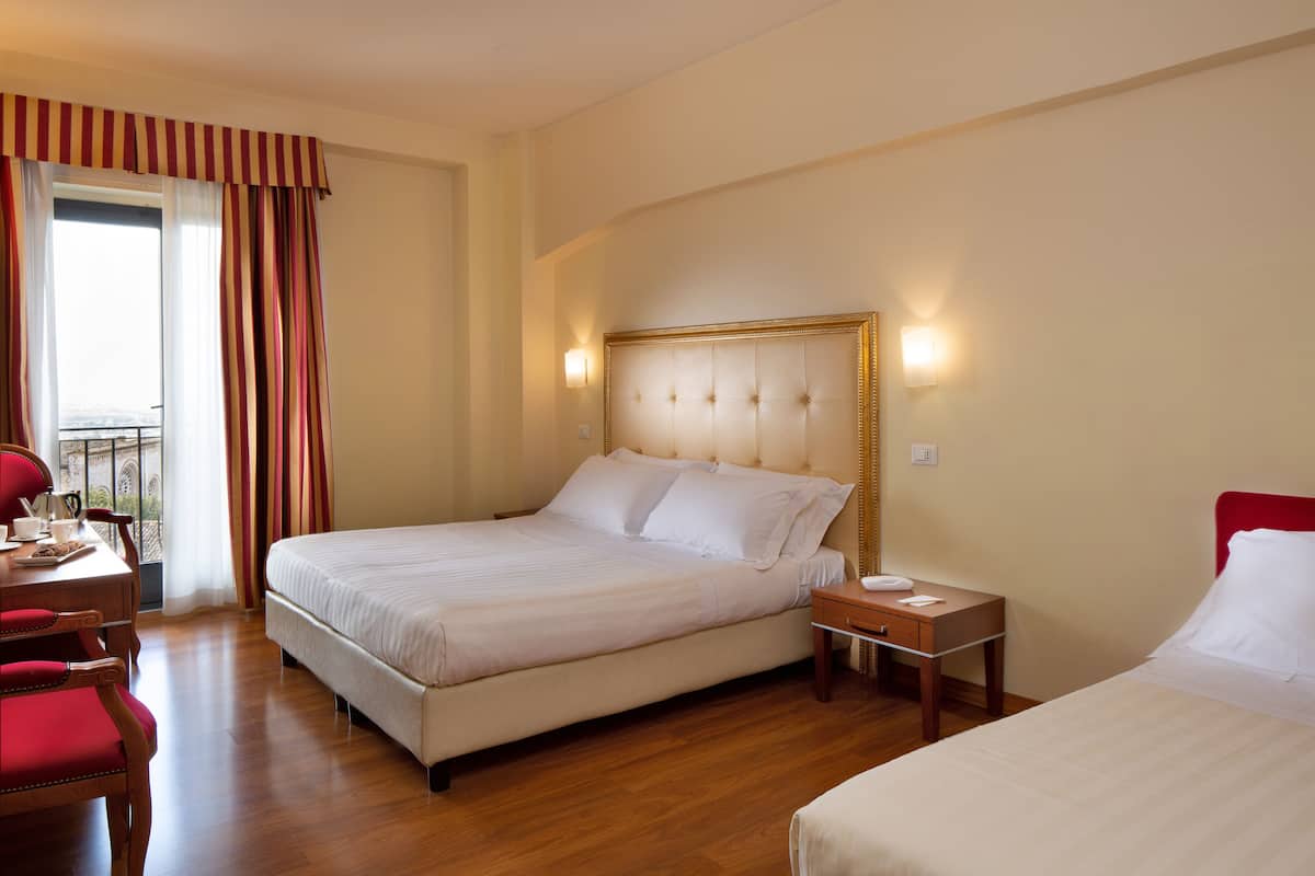 A hotel room with a large bed, a bedside table, a desk with a chair, and a window with curtains.