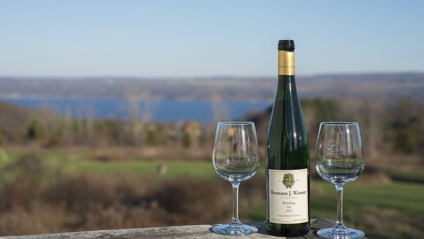 A bottle of Hermann J. Wiemer Vineyard Riesling 2014 wine with two empty glasses on a wooden surface, set against a scenic background. - Savannah House Wine Country Inn & Cottages (Himrod)