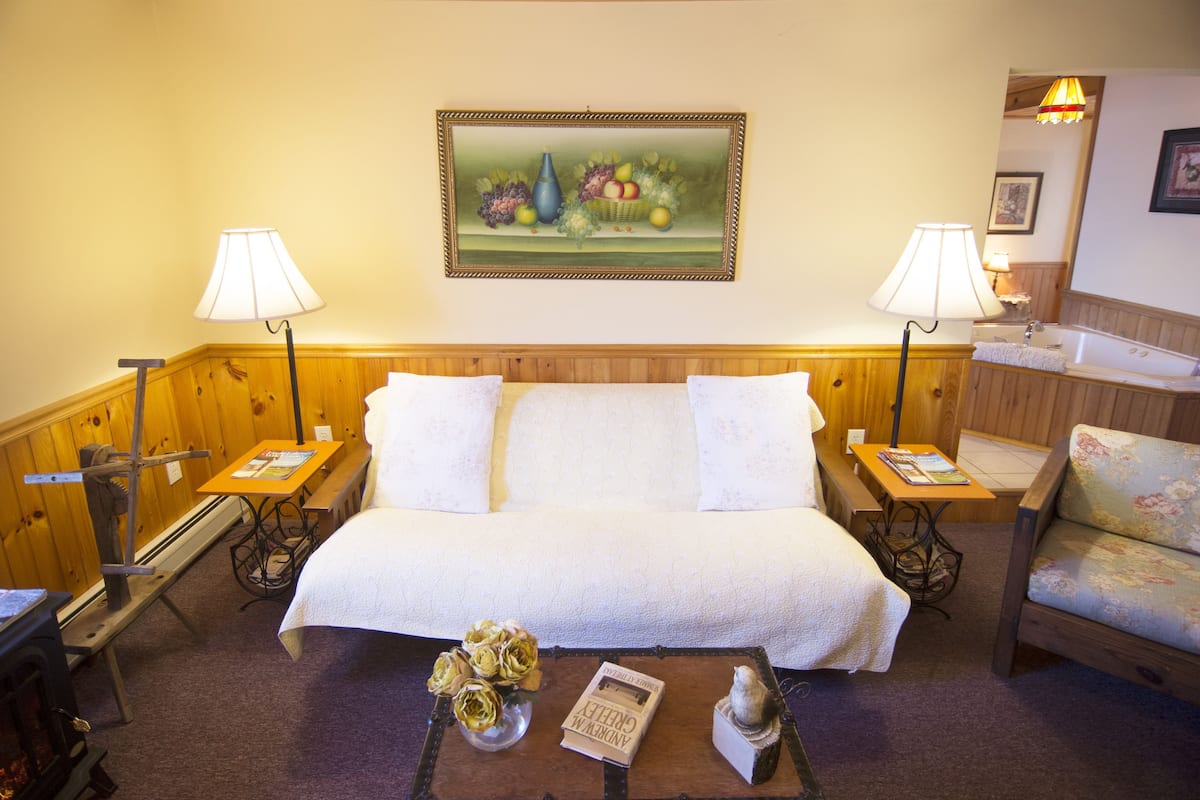 Una habitación acogedora con una cama, dos mesitas de noche con lámparas, un banco de madera, una pequeña mesa con un libro y flores, y un cuadro en la pared.