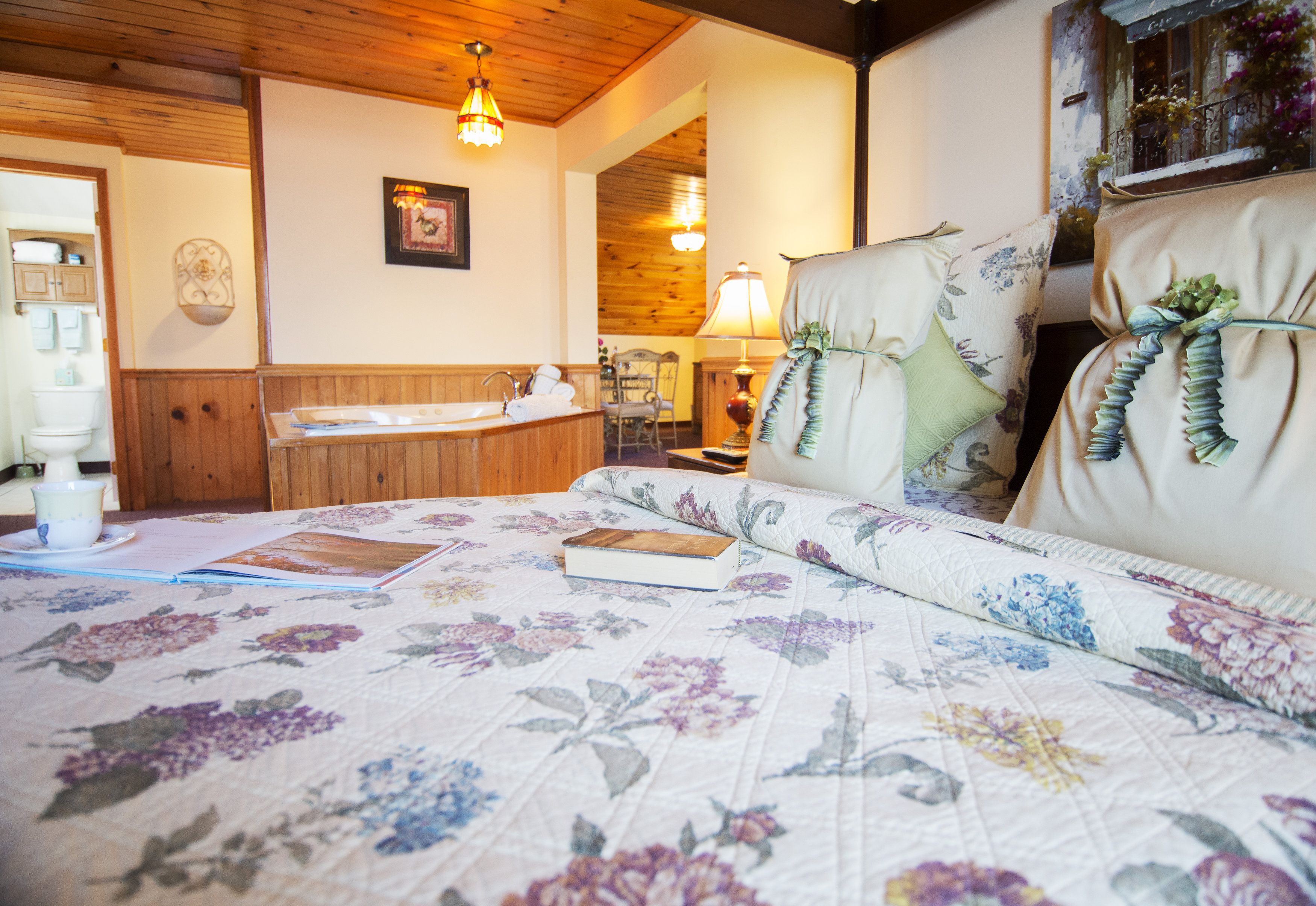 A bedroom with a floral bedspread, a wooden headboard, a bedside table with a lamp, and a view of the bathroom.