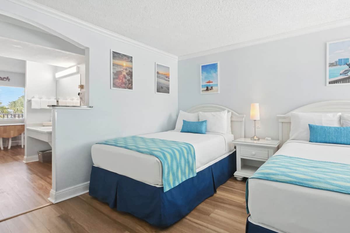 A hotel room with two beds, a desk, and a chair. There are framed pictures on the walls and a view of the beach through the window.