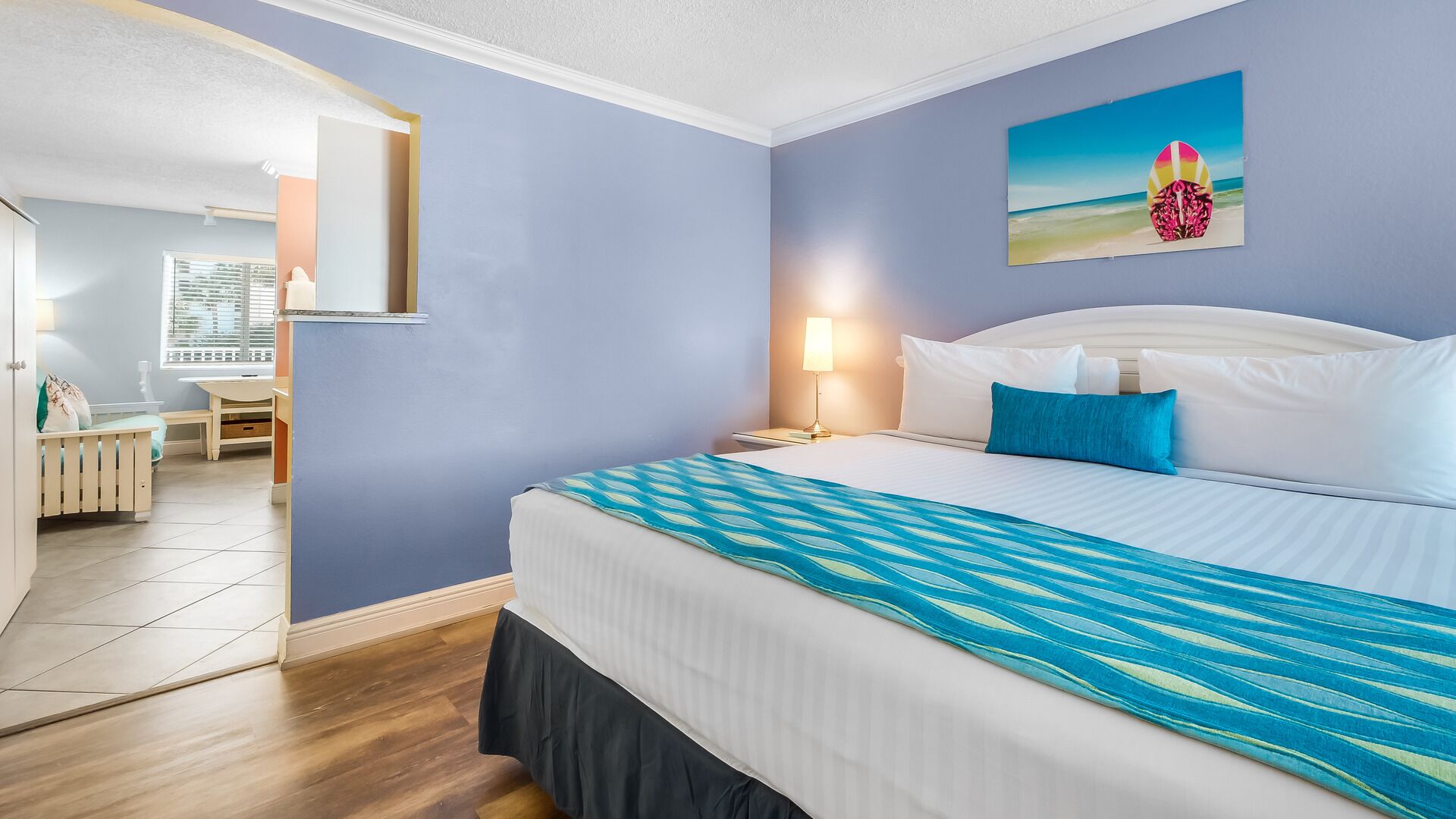 A hotel room with a bed, a nightstand, a lamp, a painting of a beach scene, and a view of the room's interior.