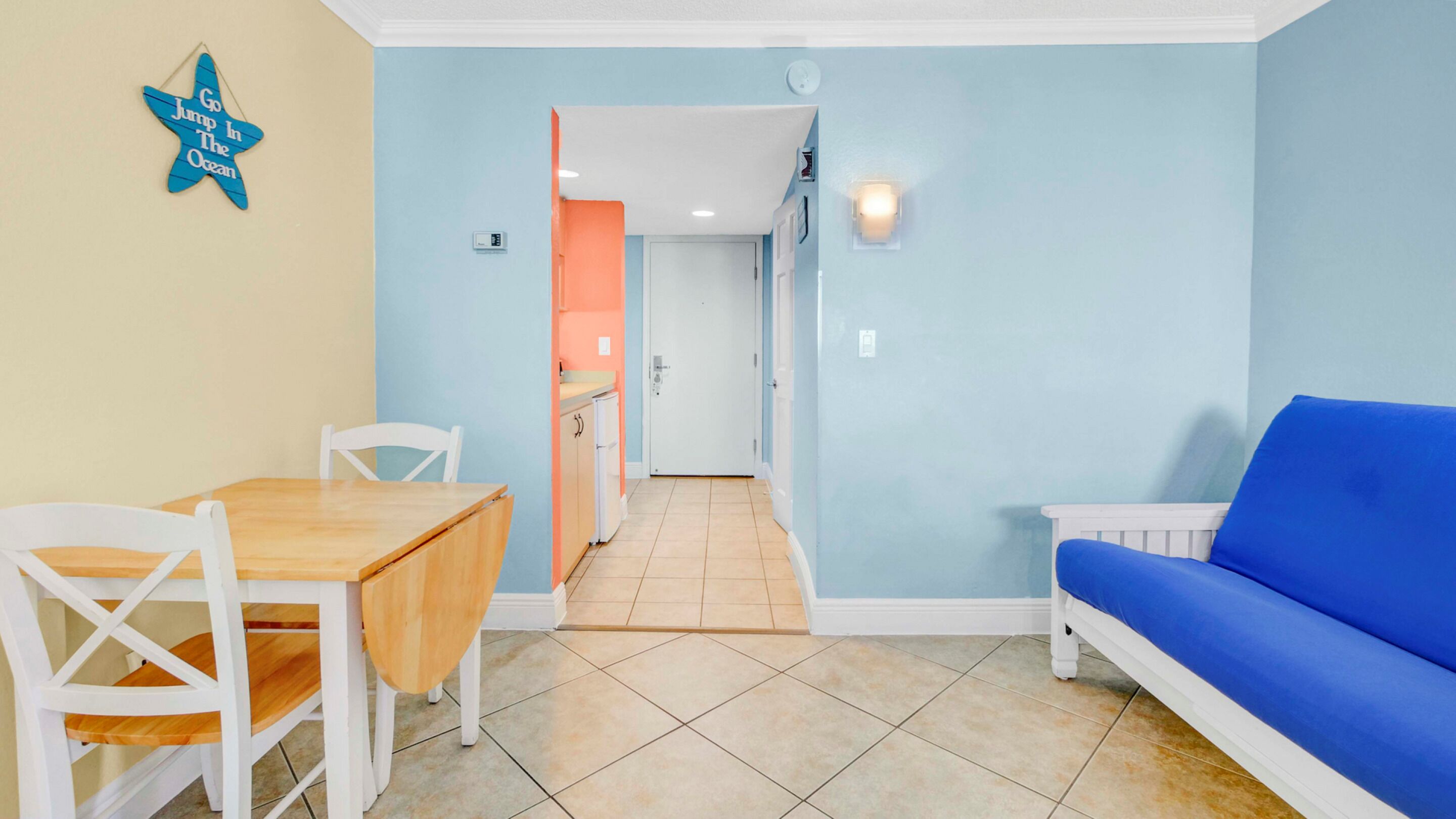 A room with a dining table, chairs, a blue sofa, and a kitchen area in the background.