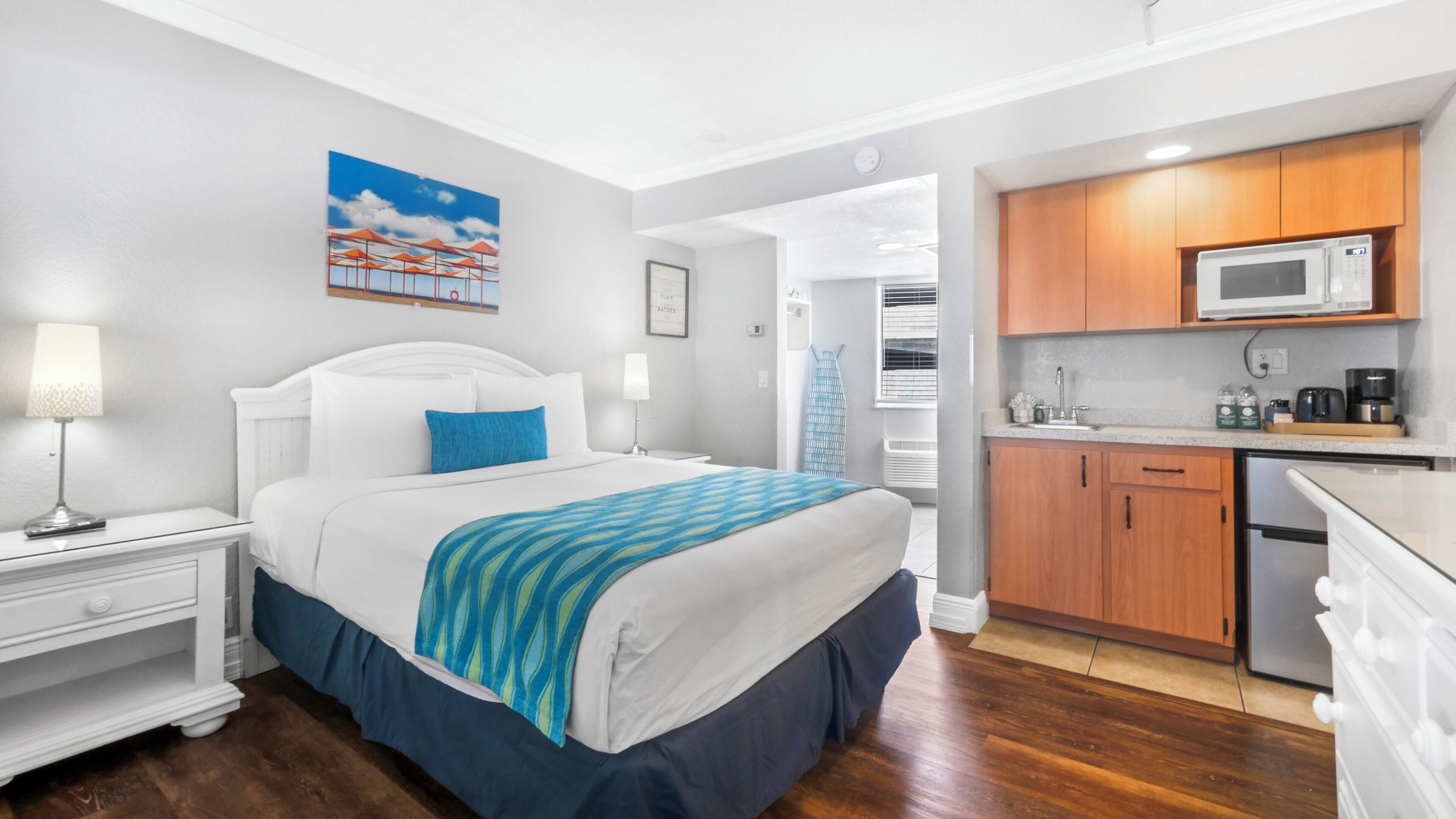 A hotel room with a bed, a nightstand, a lamp, a painting, a kitchenette with a microwave, and a refrigerator.