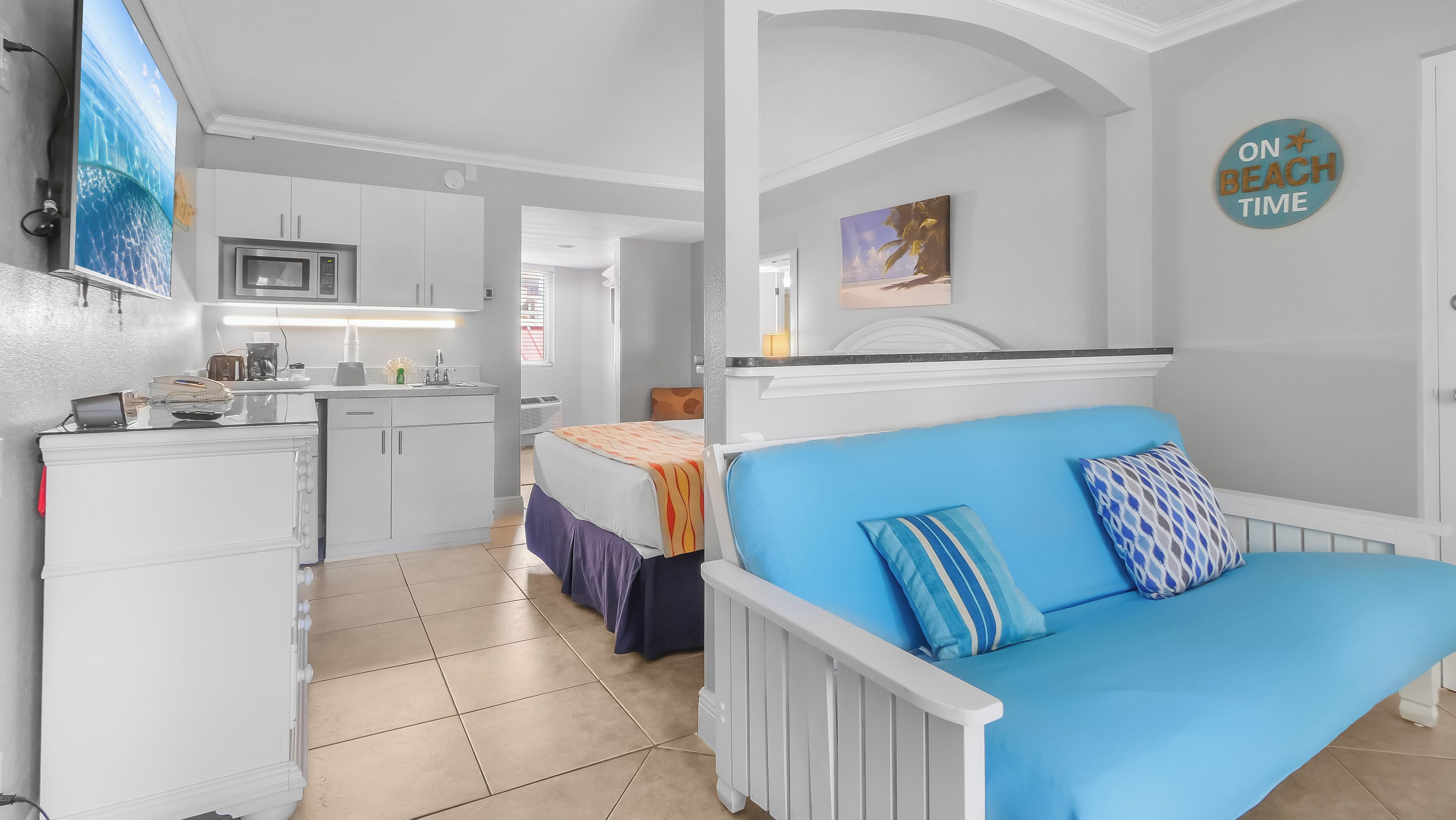 A compact hotel room with a kitchenette, a blue sofa, and a 'On Beach Time' sign.