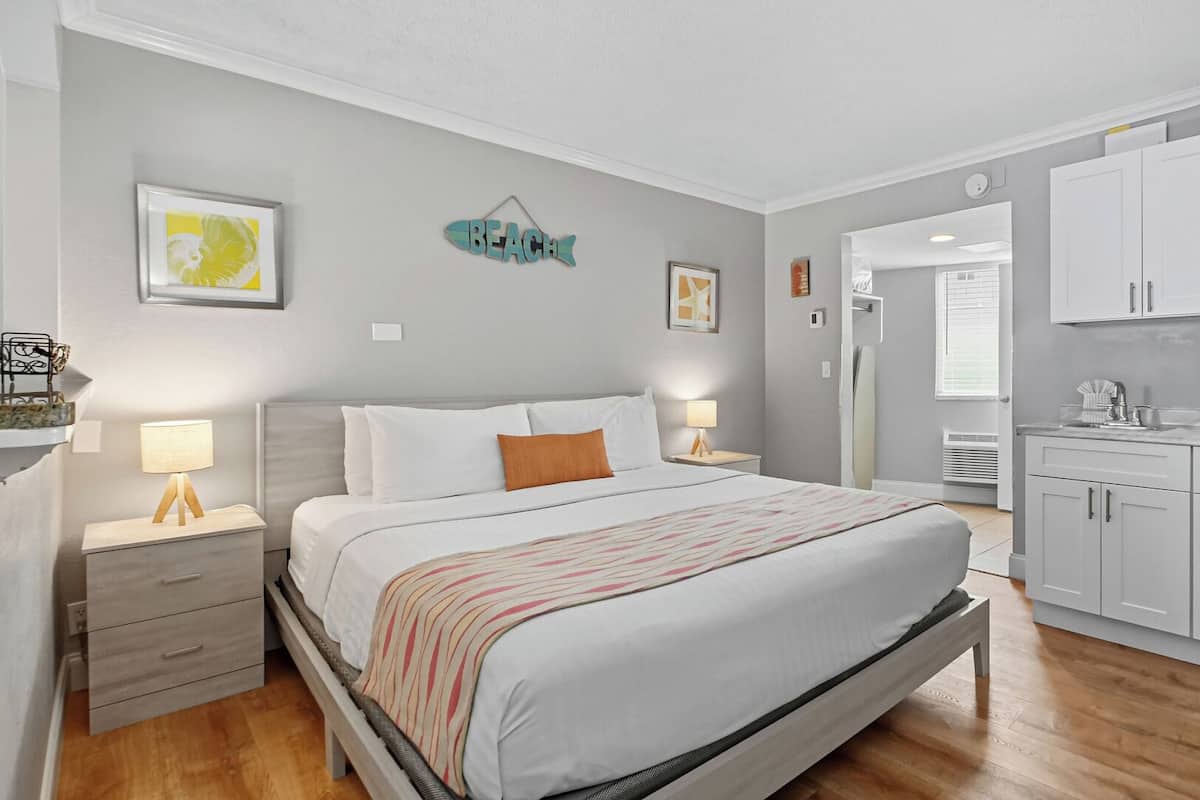 A modern bedroom with a large bed, bedside tables, and a small desk. There is a kitchenette with a microwave and cabinets.