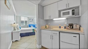 A compact hotel room with a kitchenette, a bed with blue bedding, a window with blinds, and a wall-mounted TV.
