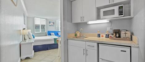 Habitación de hotel compacta con kitchenette, cama con ropa de cama azul, ventana con persianas y televisor montado en la pared.