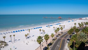 A beach with a long stretch of white sand, palm trees, and a pier in the background.