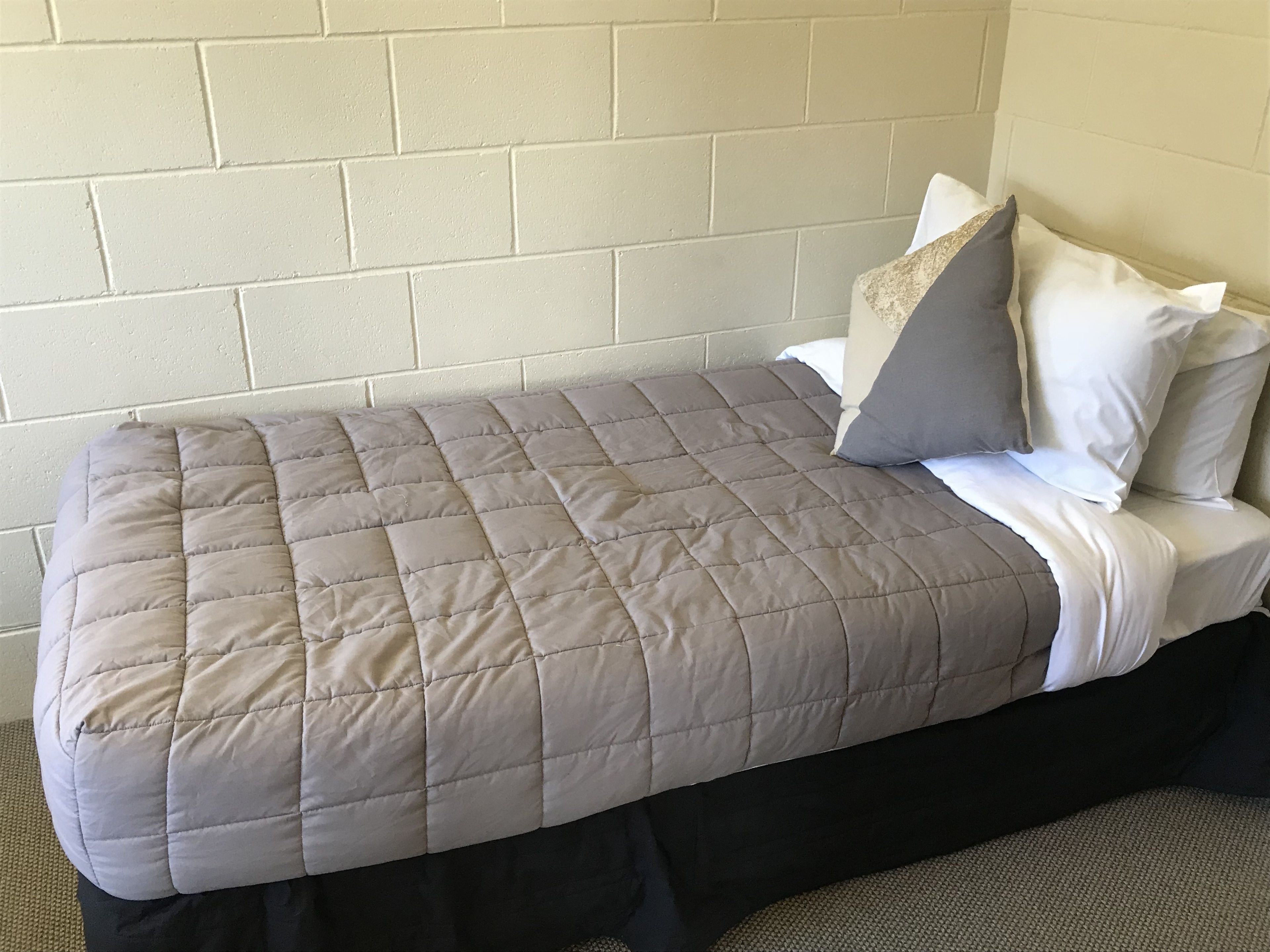 Un lit simple avec une couvre-lit matelassée grise, des oreillers blancs et une jupe de lit de couleur foncée.