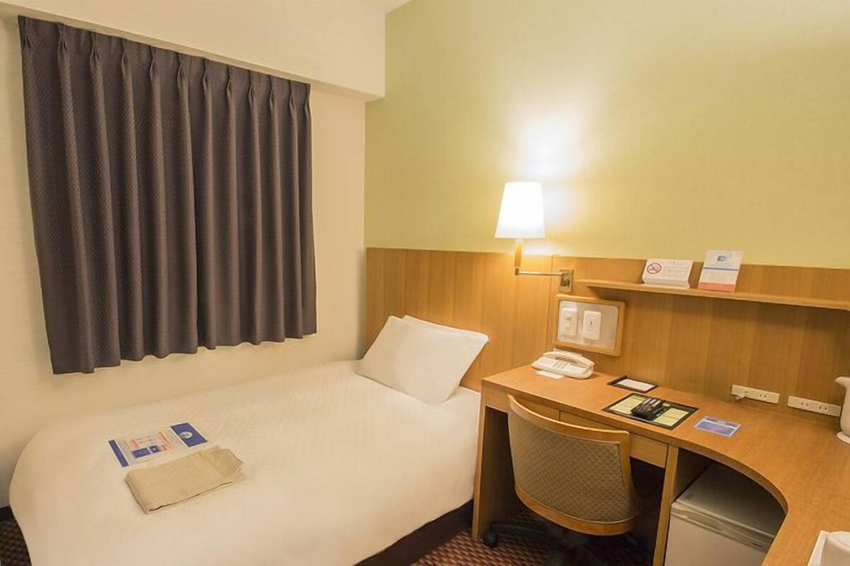 A hotel room with a bed, a desk, a chair, a television, a lamp, and a wall-mounted shelf with items.