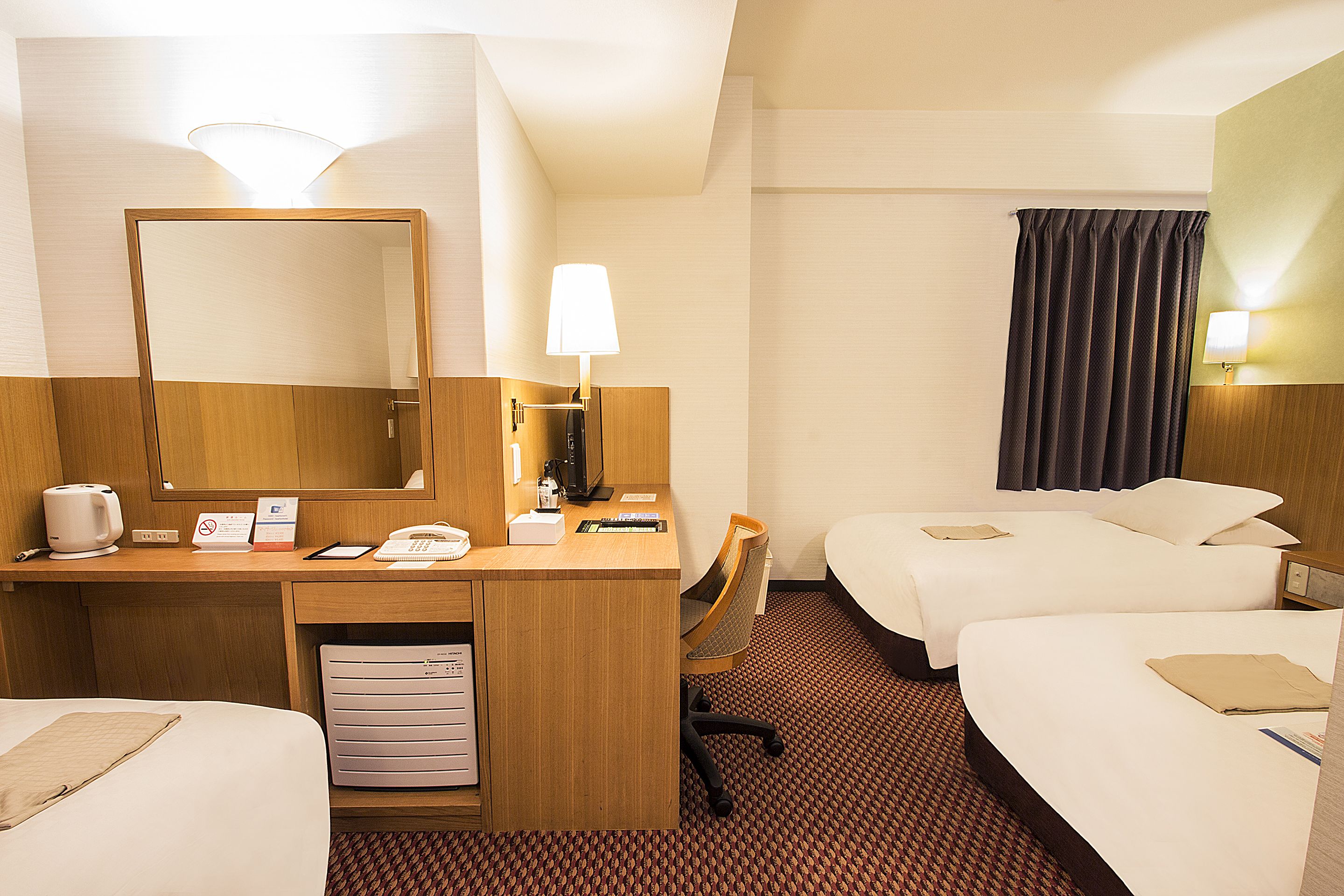 Habitación de hotel con escritorio de madera, dos camas, una silla, un espejo, una lámpara, una mesita de noche y piso alfombrado.