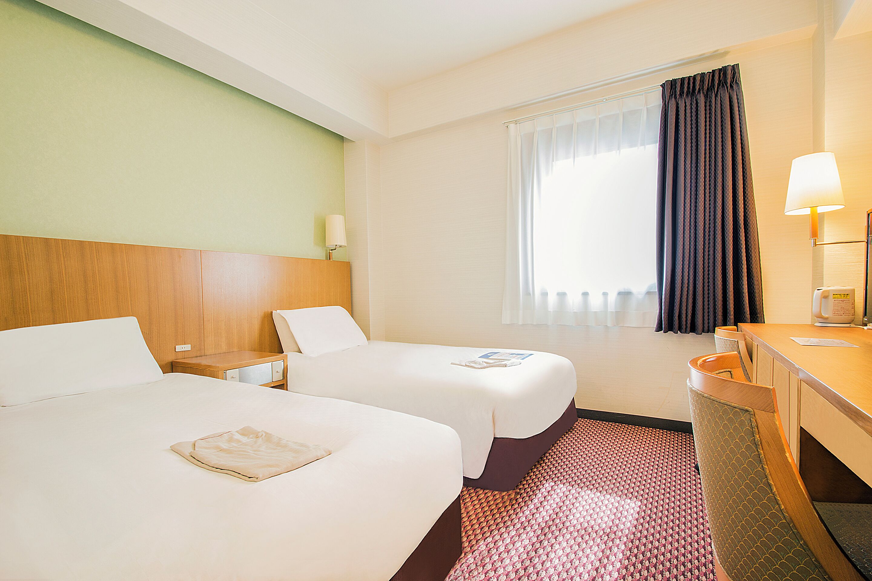 A hotel room with two beds, a desk, a chair, a window with curtains, and a lamp.