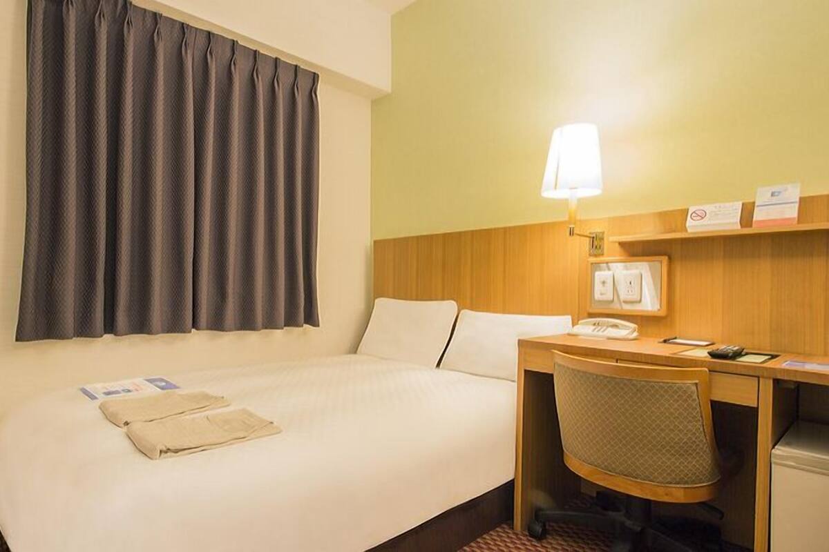 A hotel room with a bed, a desk with a lamp, a telephone, and a small shelf with items.