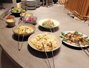 Uma mesa de buffet com diversos pratos, incluindo saladas, legumes e carnes, com utensílios de servir.