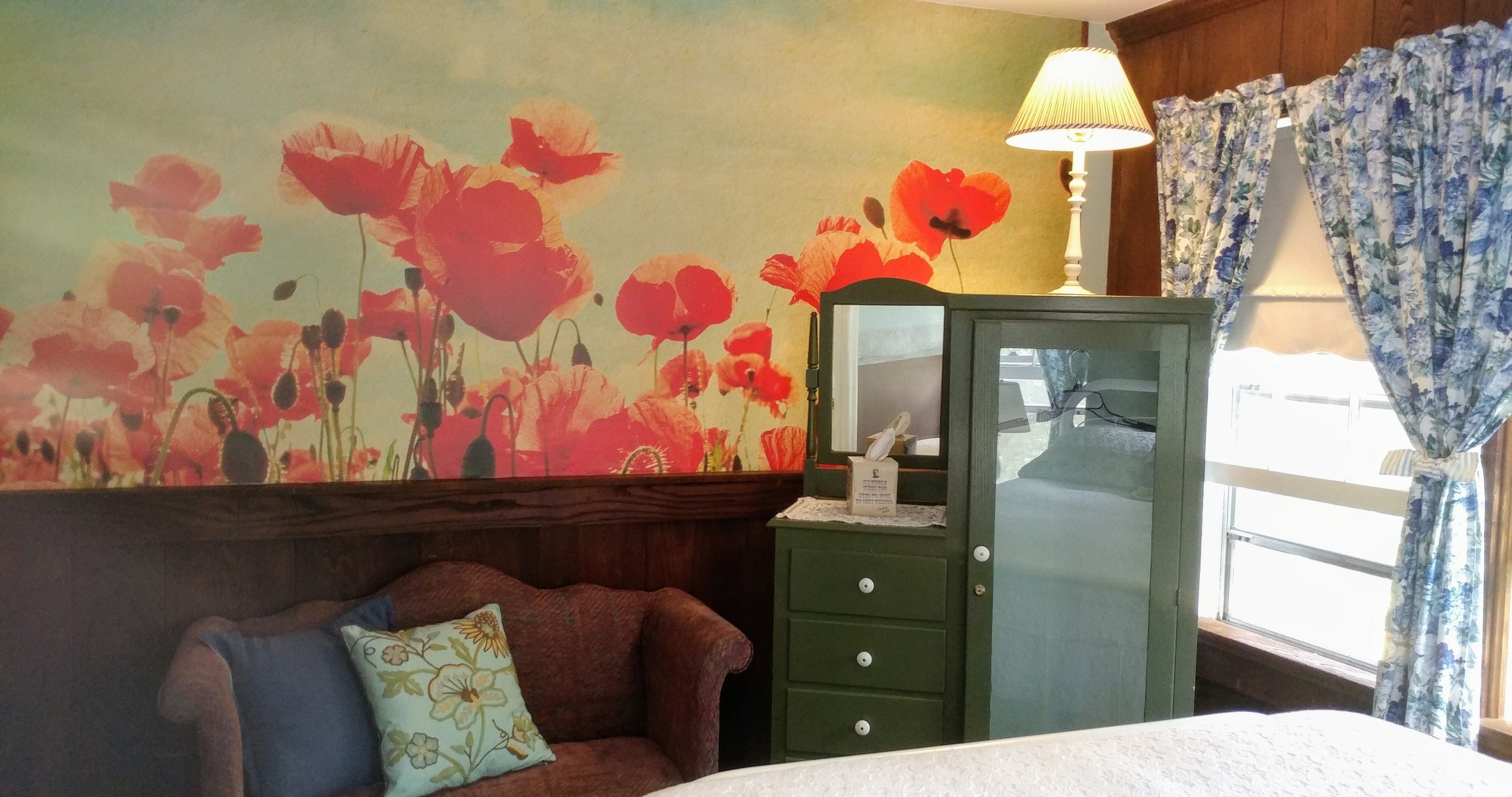 Une pièce avec un mur décoré d’un motif floral, un meuble vert, un fauteuil bleu et une fenêtre avec des rideaux.