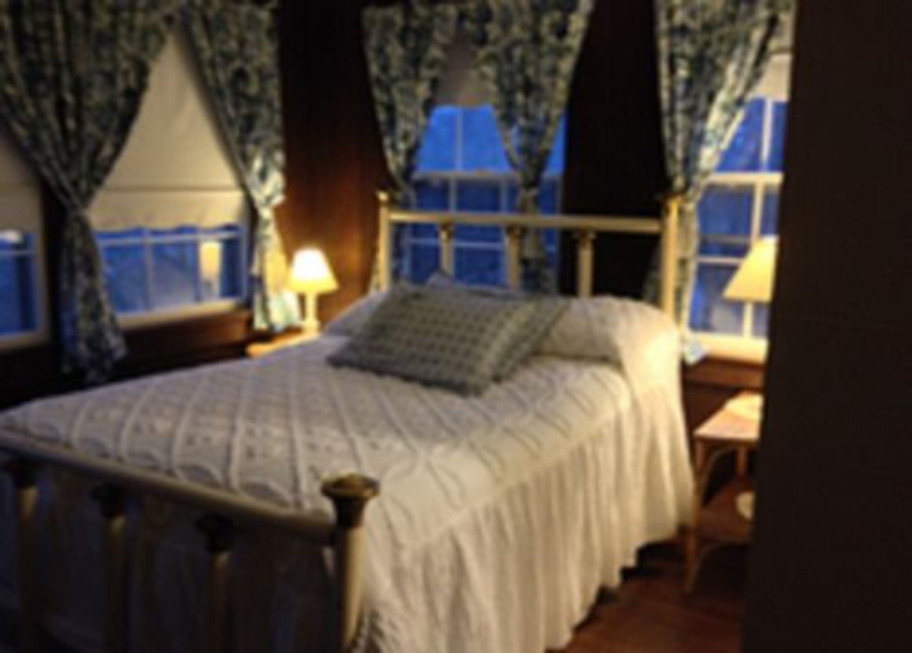 Un dormitorio con cama, cortinas, una ventana y una mesita de noche con lámpara.