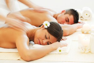 A spa setting with two individuals lying on a massage table, a white flower in hair, and candles on the side.