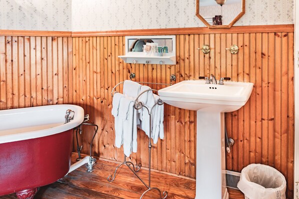 A bathroom with a wooden panel wall, a red clawfoot bathtub, a white pedestal sink, and a mirror above the sink.