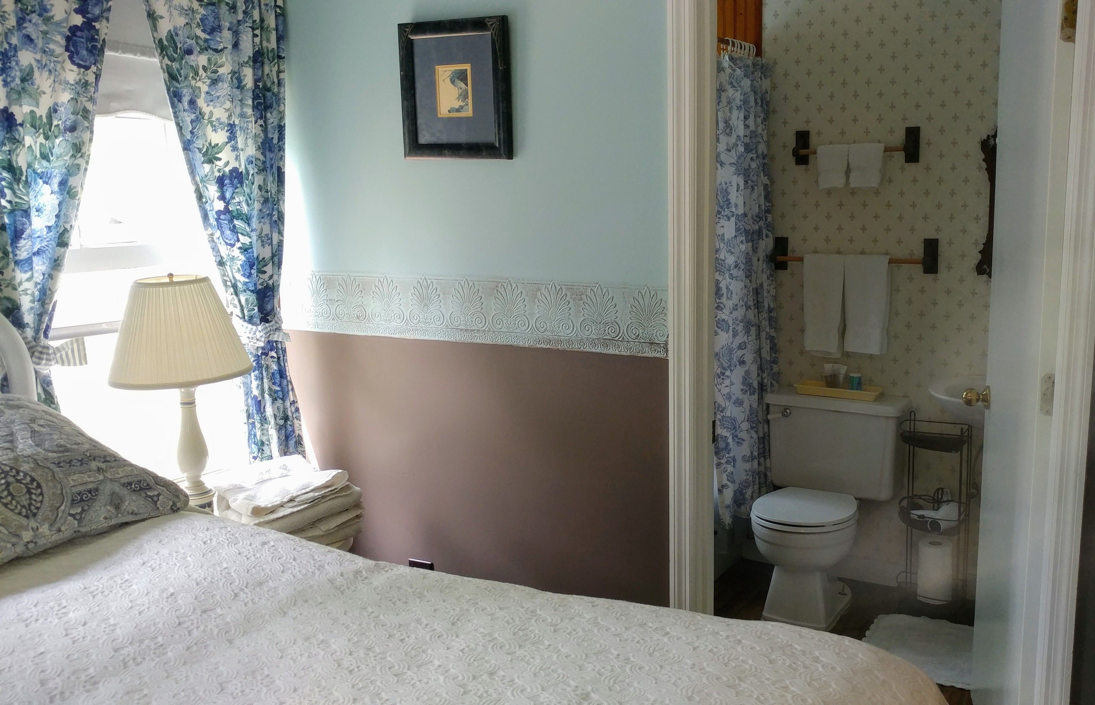 A bedroom with a bed, a bedside lamp, a window with floral curtains, and a bathroom with a toilet, sink, and towel rack.