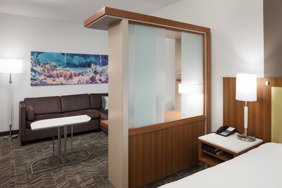 A hotel room with a sofa, a small table, a mirror, a bedside table with a lamp, and a painting on the wall.