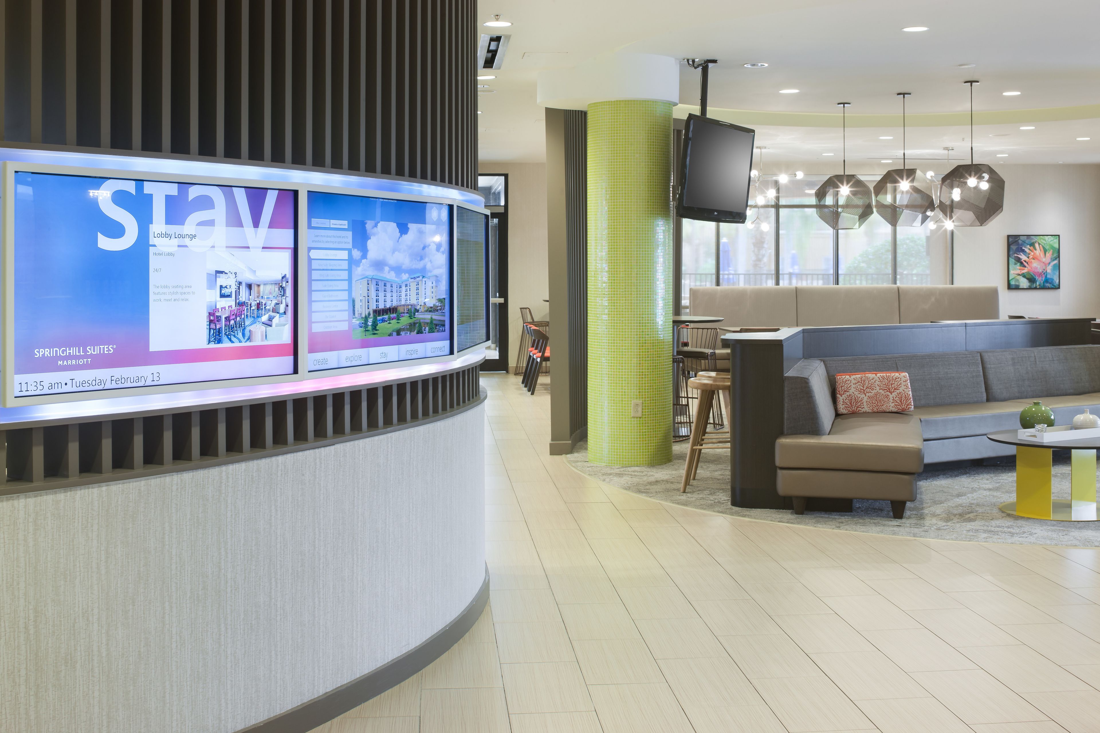 A hotel reception area with a curved reception desk, a large digital display, a seating area with a sofa and chairs, and a large window with natural light.