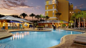Una piscina con sillones de descanso y un bar, rodeada de palmeras y un edificio de hotel.