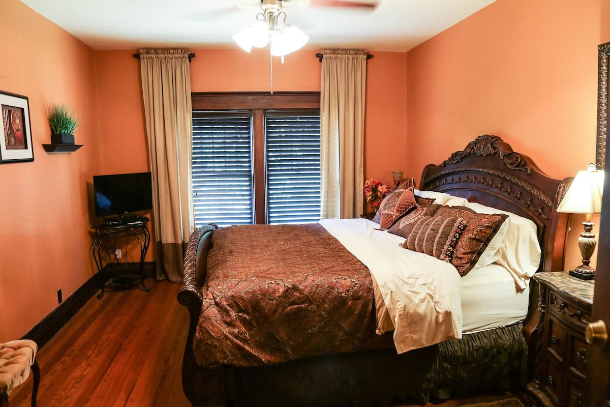 A bedroom with a large bed, a television, a ceiling fan, and a window with blinds.
