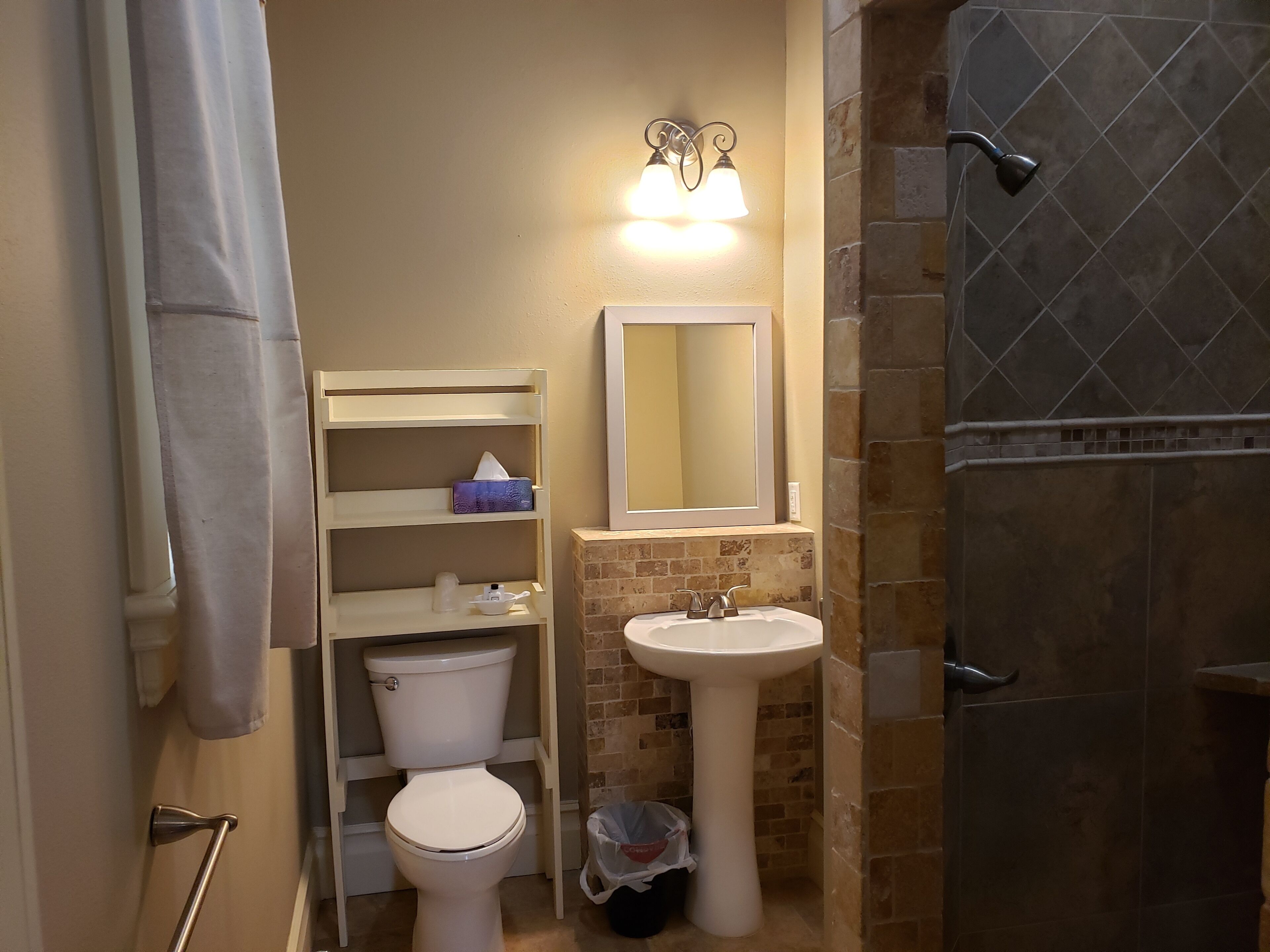 Baño pequeño y funcional con inodoro, lavabo y ducha. Hay un estante con un dispensador de pañuelos y un cesto de basura.