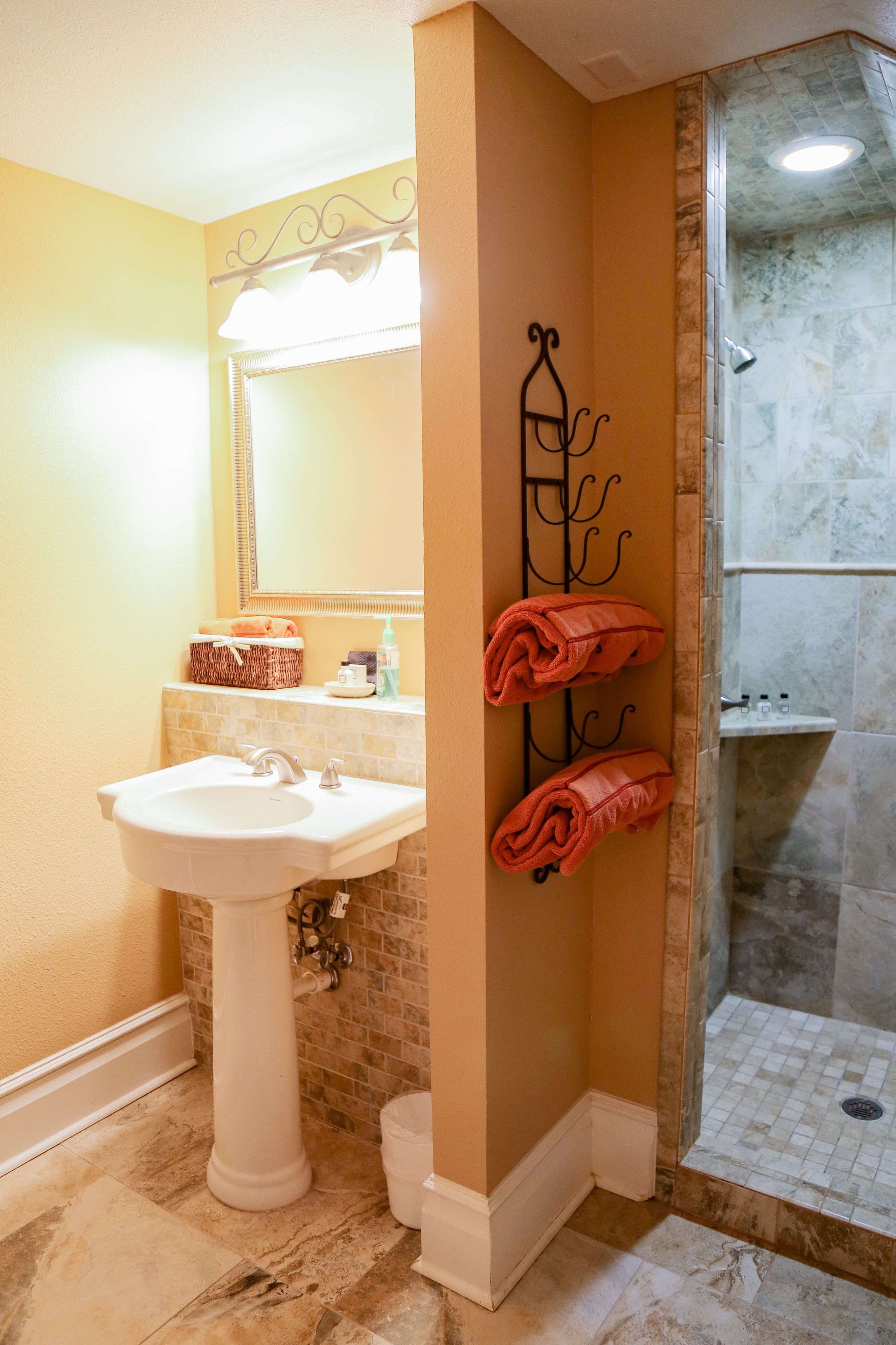 A bathroom with a pedestal sink, a mirror, a towel rack with towels, a shower area, and a tiled floor.