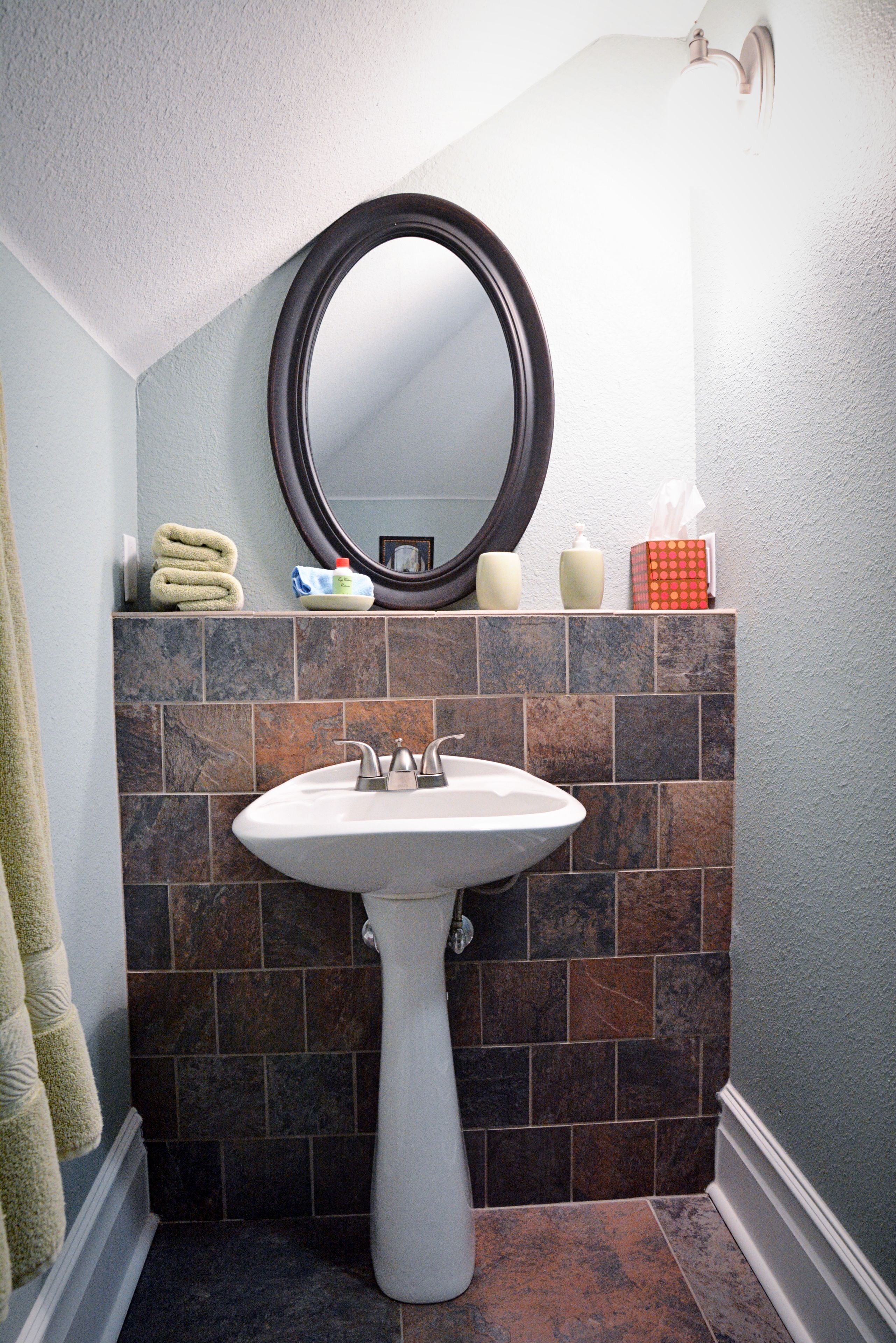 A bathroom with a white pedestal sink, dark brown and gray tiles, a round mirror, and a towel rack with folded towels.