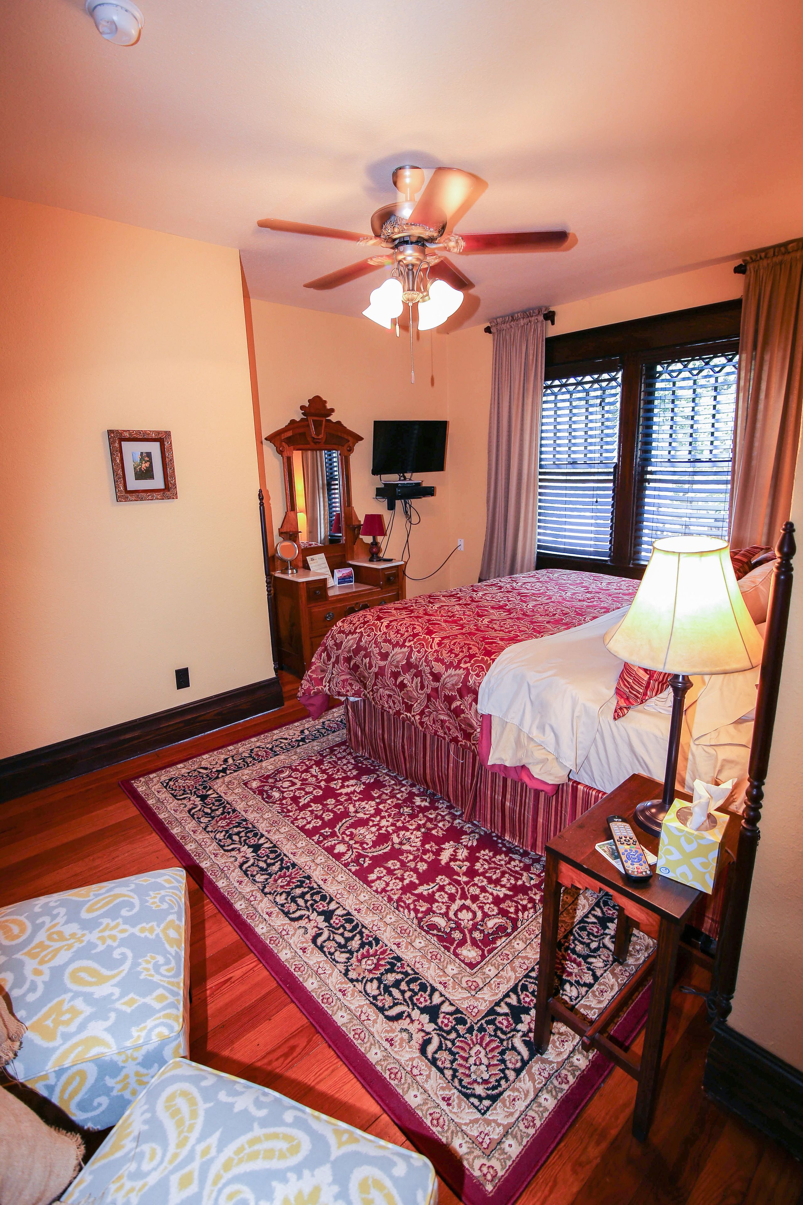 A bedroom with a bed, a ceiling fan, a nightstand, a lamp, a mirror, a dresser, a TV, and a window with curtains.