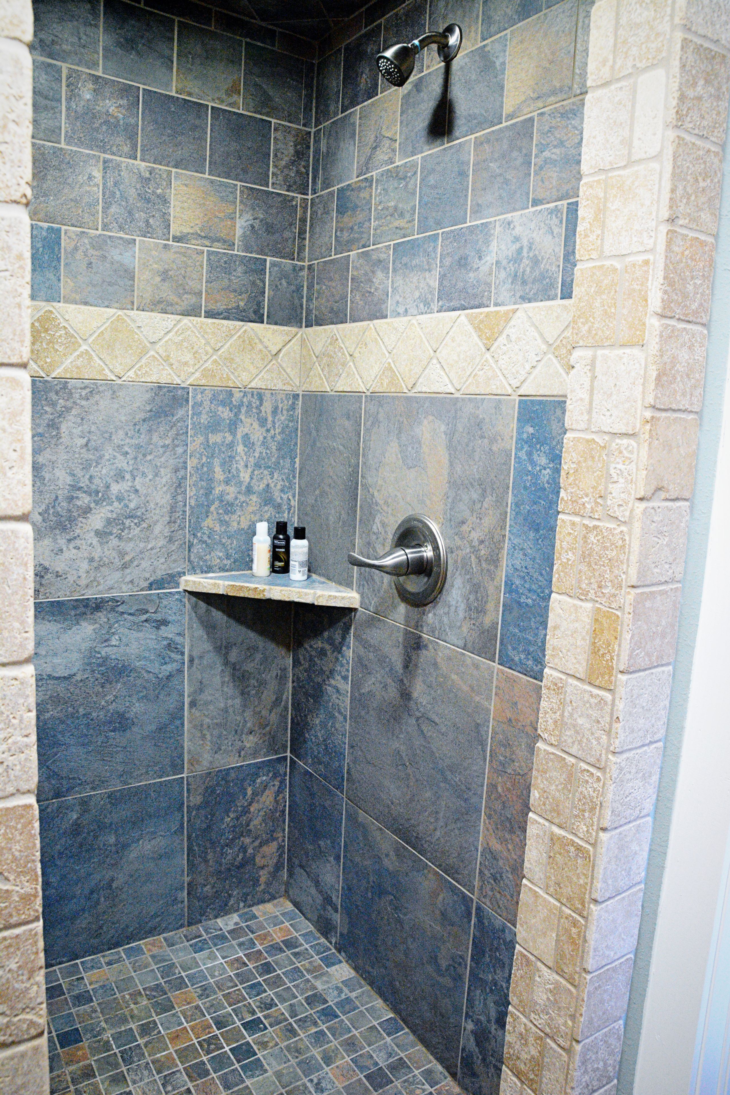 A shower area with blue and beige tiles, a showerhead, and a shelf with toiletries.