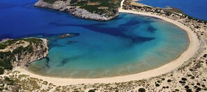 Vue aérienne d’une zone côtière avec une plage de sable, des eaux d’un bleu limpide et des affleurements rocheux.