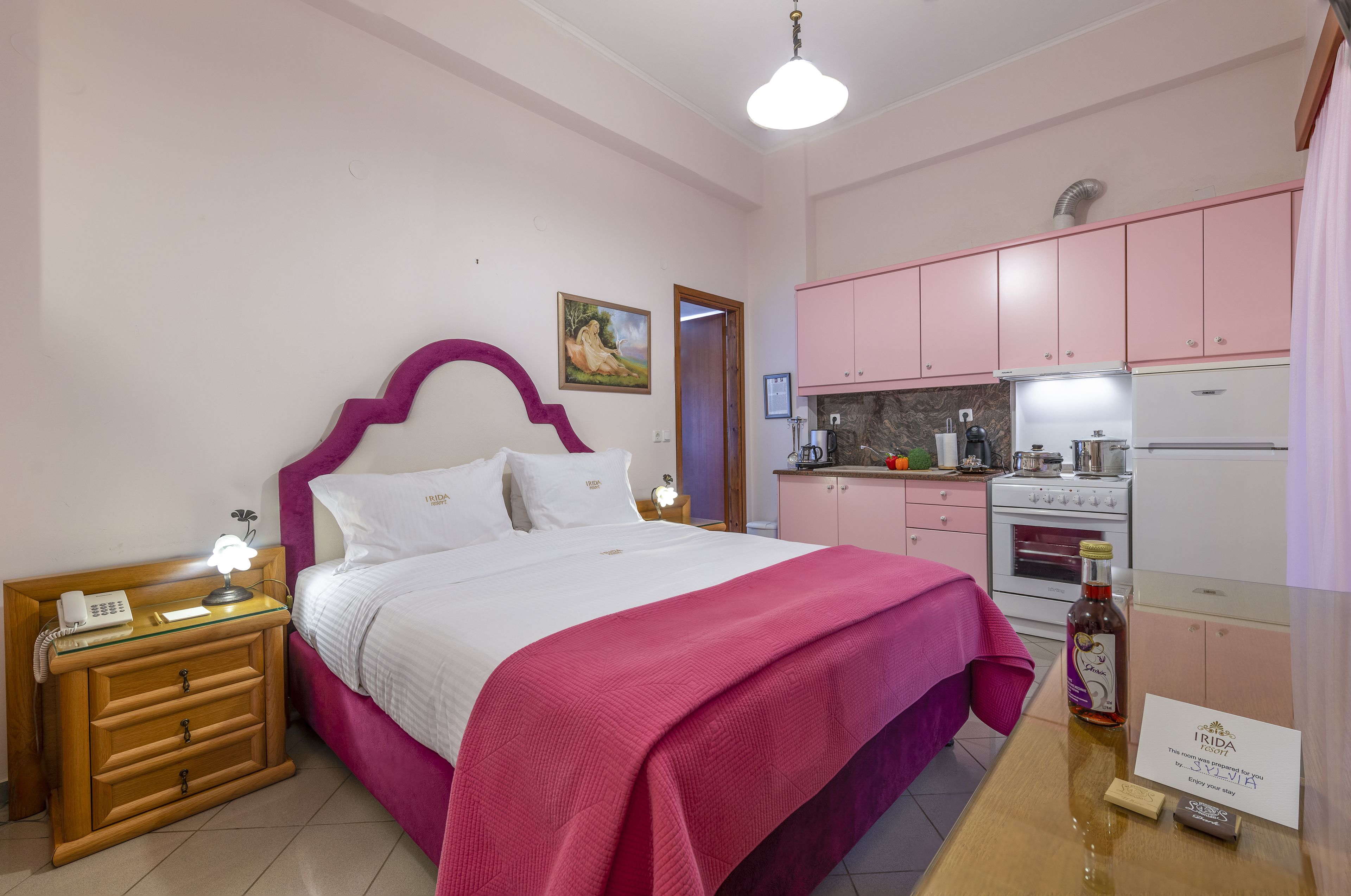 A bedroom with a bed, bedside table, and a small kitchenette area with pink cabinets and a stove.