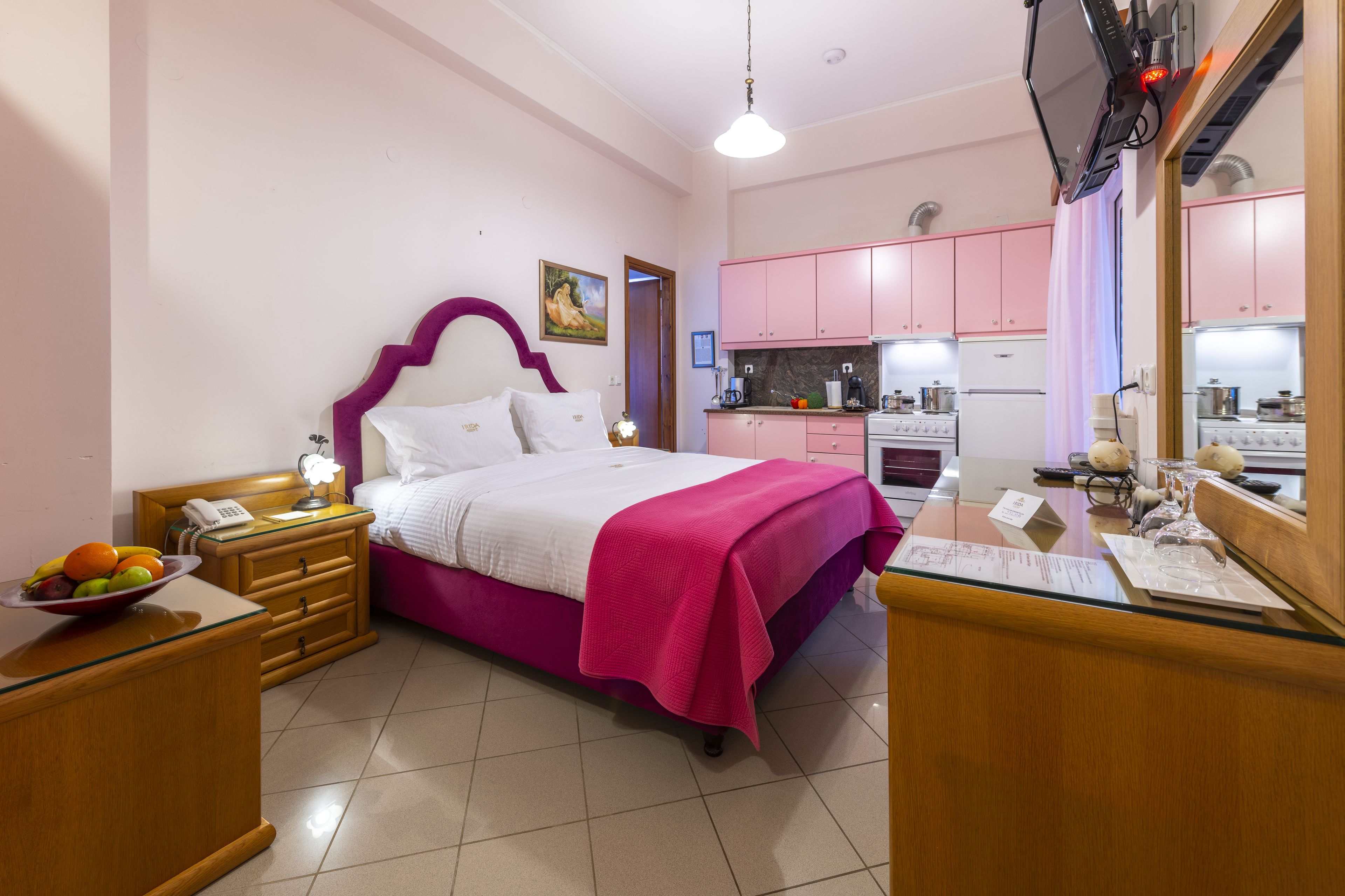 A bedroom with a bed, bedside table, and a small kitchen area with pink cabinets and a stove.