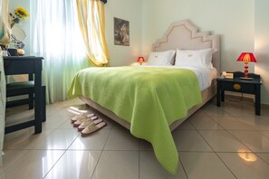 A neatly made bed with a green bedspread, a nightstand with a red lamp, and a chair with slippers.