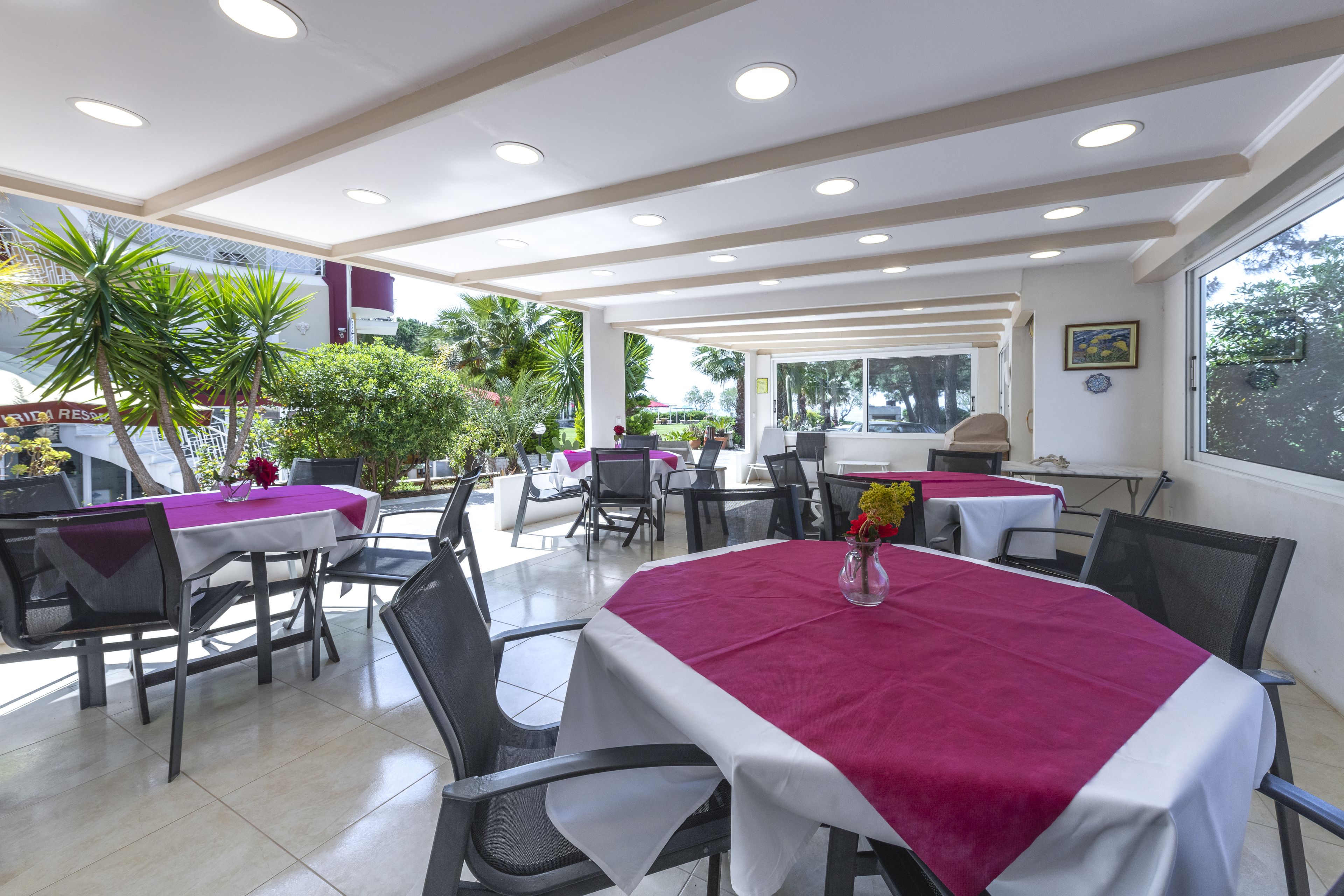 A restaurant with outdoor seating, white tables with red tablecloths, and a view of greenery outside.