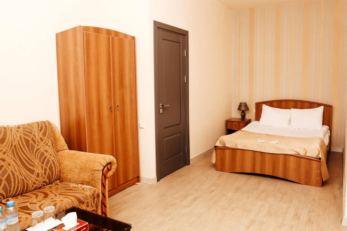 A hotel room with a wooden wardrobe, a bed with a wooden headboard, a bedside table with a lamp, and a patterned sofa.