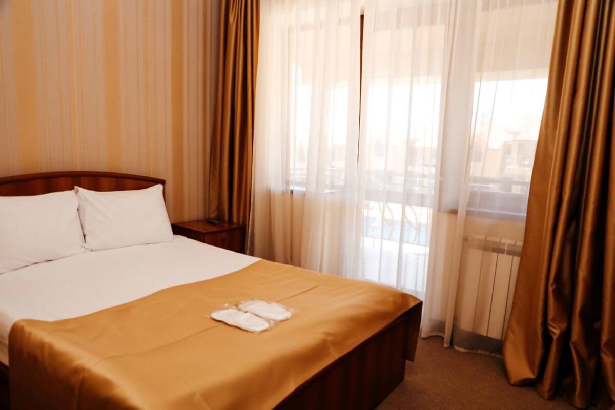 A hotel room with a large bed, a nightstand, and a view through sheer curtains.