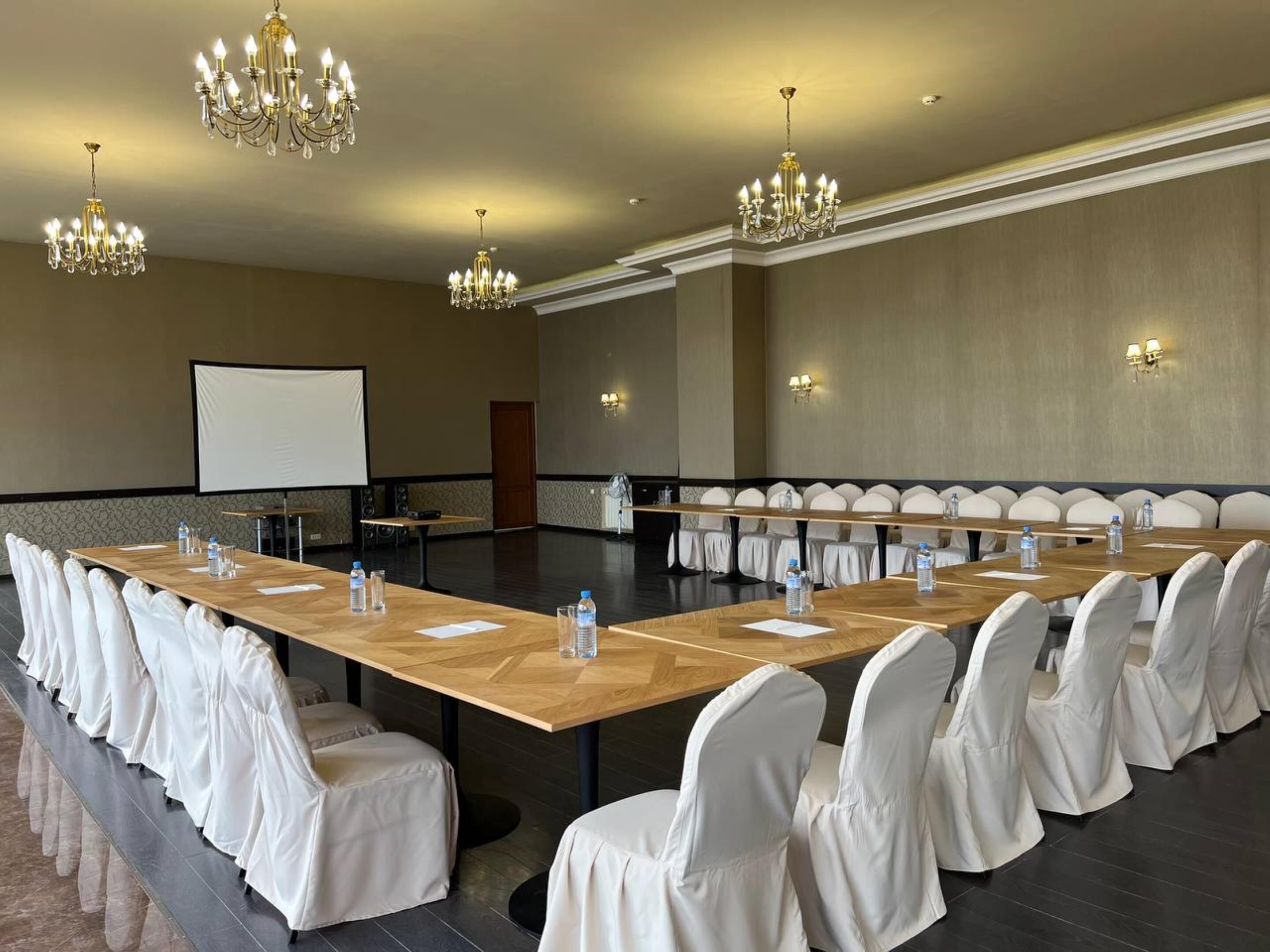 A conference room with a long table, white chairs, chandeliers, and a projector screen.