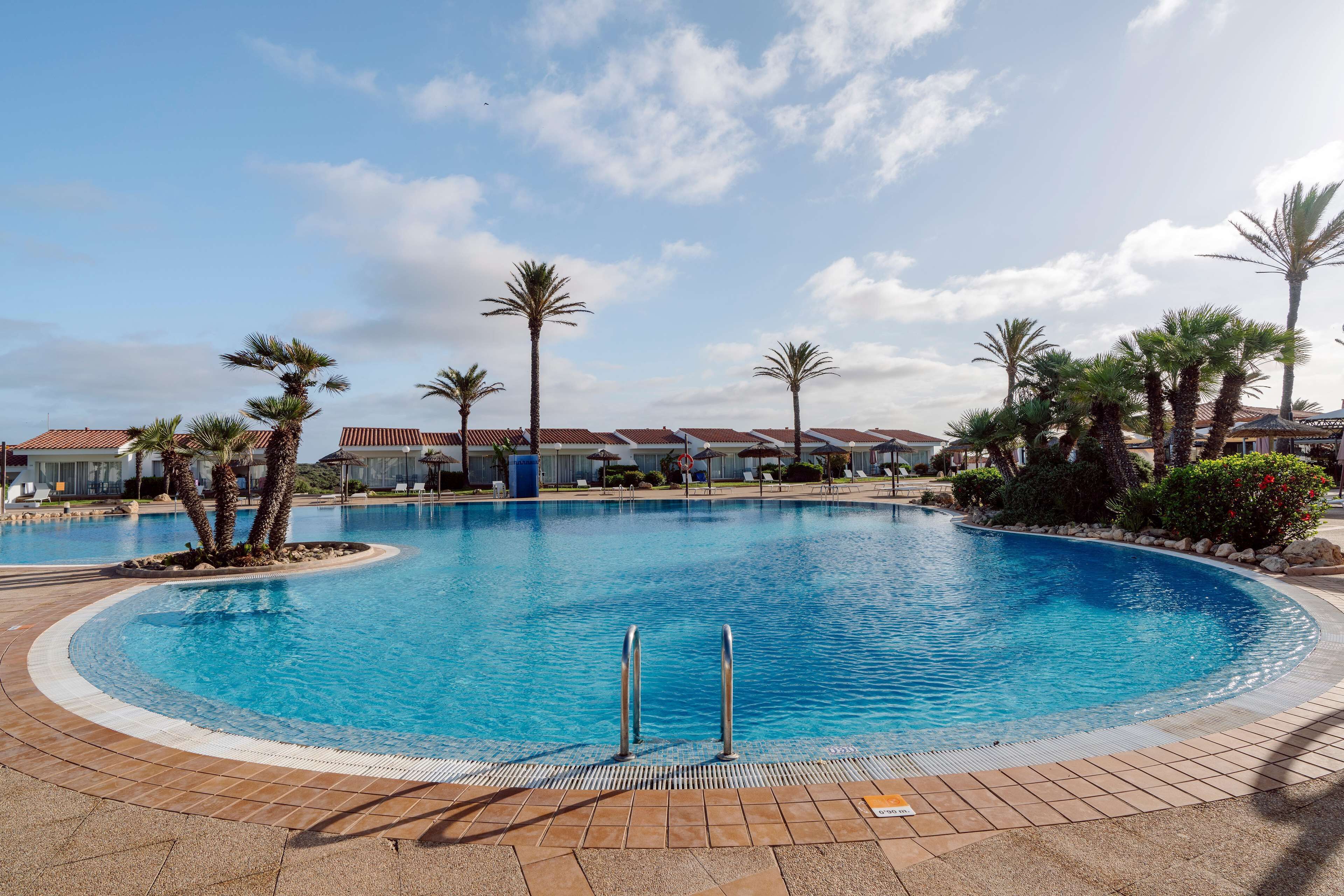 Una piscina con palme e un solarium piastrellato, circondata da edifici con tetti rossi.