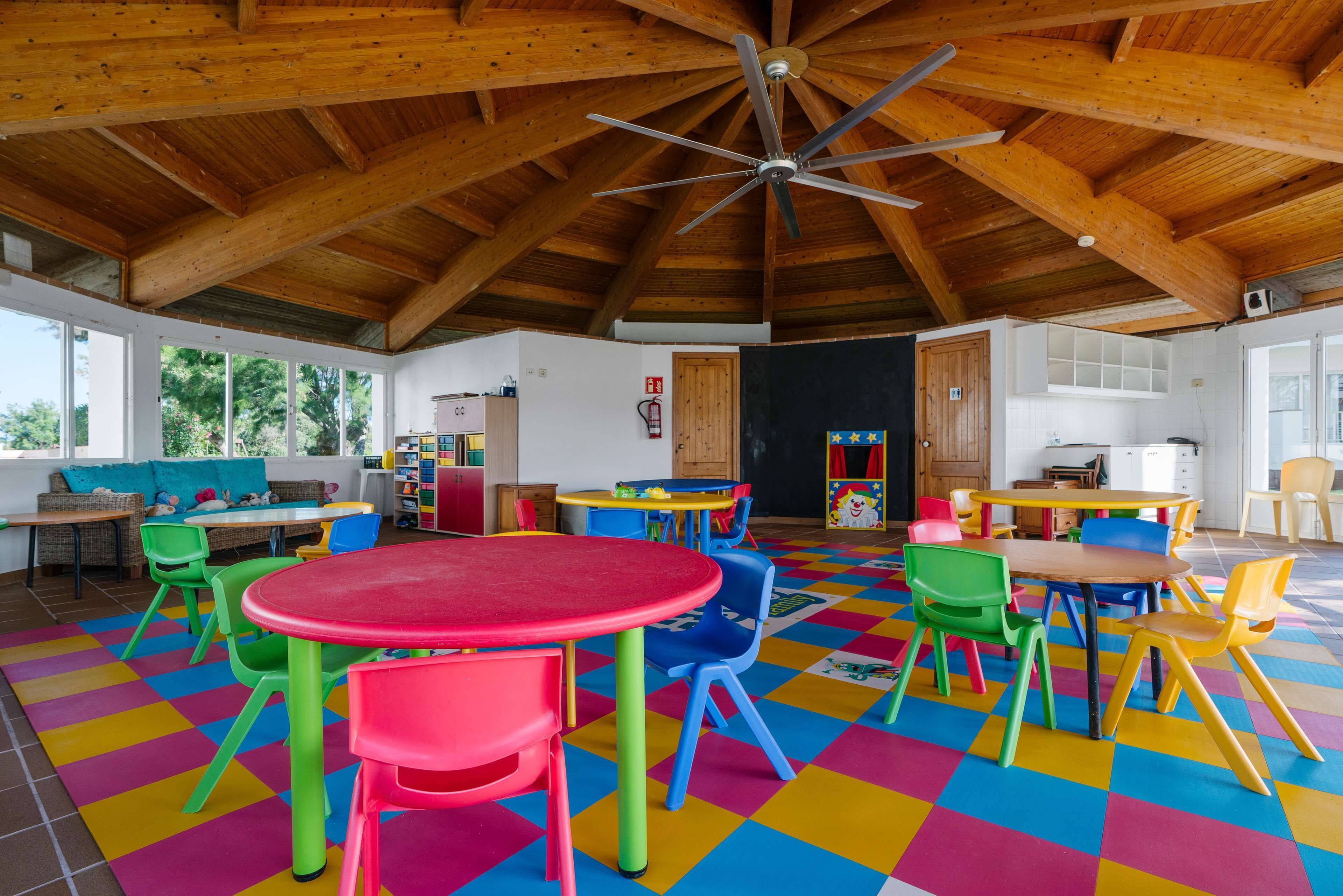 Ein geräumiges Zimmer mit Holzbalken, einem Deckenventilator und farbig kariertem Boden. Tische und Stühle sind für Essen oder andere Aktivitäten angeordnet.